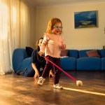 Two-year-old Martha Thompson, in turquoise glasses, walking across a wooden floor with a pink cane, her mum smiling behind her