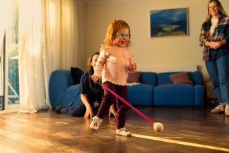 Two-year-old Martha Thompson, in turquoise glasses, walking across a wooden floor with a pink cane, her mum smiling behind her