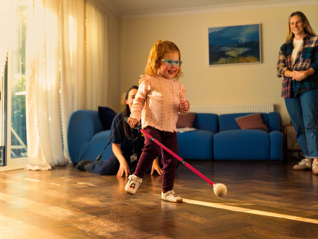 Two-year-old Martha Thompson, in turquoise glasses, walking across a wooden floor with a pink cane, her mum smiling behind her