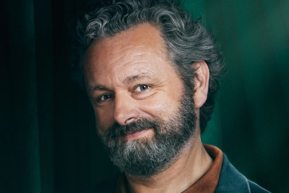 Michael Sheen smiling in a portrait for The Celebrity Traitors, grey curly hair and beard against a green curtain
