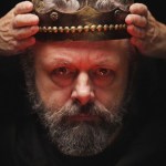 Michael Sheen, bearded and grey, lowers a battered gold crown onto his own head against a black background