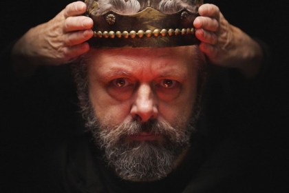 Michael Sheen, bearded and grey, lowers a battered gold crown onto his own head against a black background
