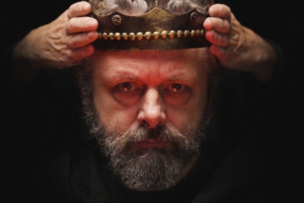Michael Sheen, bearded and grey, lowers a battered gold crown onto his own head against a black background