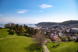 A panoramic view over Mumbles from the park above Oystermouth, with the village, beach, pier and lighthouse stretching along the curve of Swansea Bay under a blue sky