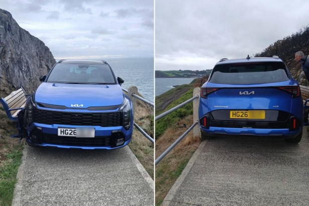 A blue Kia Sportage blocking a narrow concrete clifftop footpath, its front end against a wooden bench, with the sea beyond.