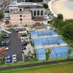 Aerial artist's impression of three blue padel courts and a landscaped car park on the former tennis courts beside the Oyster House, with Mumbles beach and the bay beyond