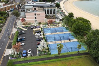 Aerial artist's impression of three blue padel courts and a landscaped car park on the former tennis courts beside the Oyster House, with Mumbles beach and the bay beyond