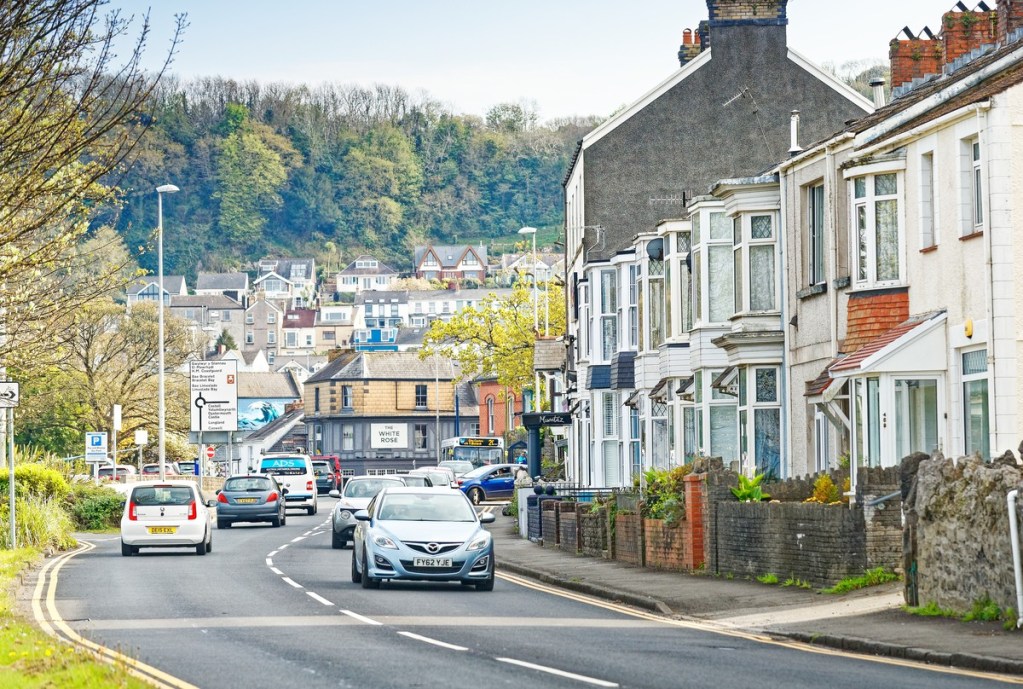 Traffic on Mumbles Road passing terraced houses and the White Rose pub, with the wooded hillside and houses of Mumbles rising behind