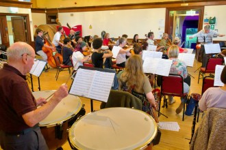 Neath Symphony Orchestra rehearsing in a wood-panelled hall, with a timpanist in the foreground, string players at music stands and the conductor at the far end