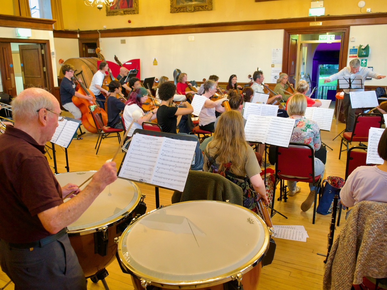 Neath Symphony Orchestra rehearsing in a wood-panelled hall, with a timpanist in the foreground, string players at music stands and the conductor at the far end