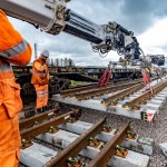 Railway engineers in orange hi-vis clothing watch as a crane lowers a new section of track with concrete sleepers into place