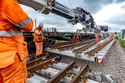 Railway engineers in orange hi-vis clothing watch as a crane lowers a new section of track with concrete sleepers into place