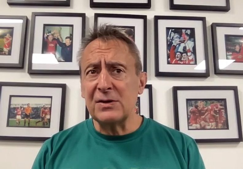 Scarlets director of rugby Nigel Davies in a green club polo shirt speaking at a press conference in front of a wall of framed rugby photographs