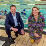 A man in a navy suit and a woman in a colourful patterned dress crouch smiling at the edge of a swimming pool while children in blue swim caps splash in the water behind them