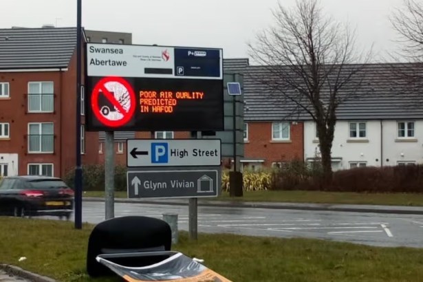 A roadside digital sign on New Cut Road, Swansea, reading "poor air quality predicted in Hafod" beside a no-vehicles symbol, with houses and a passing car behind