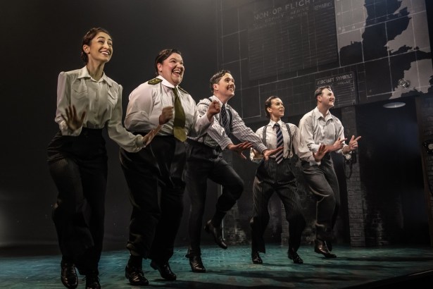 Five performers in 1940s shirts, ties and braces dancing in a line on stage in front of a wartime operations board and map