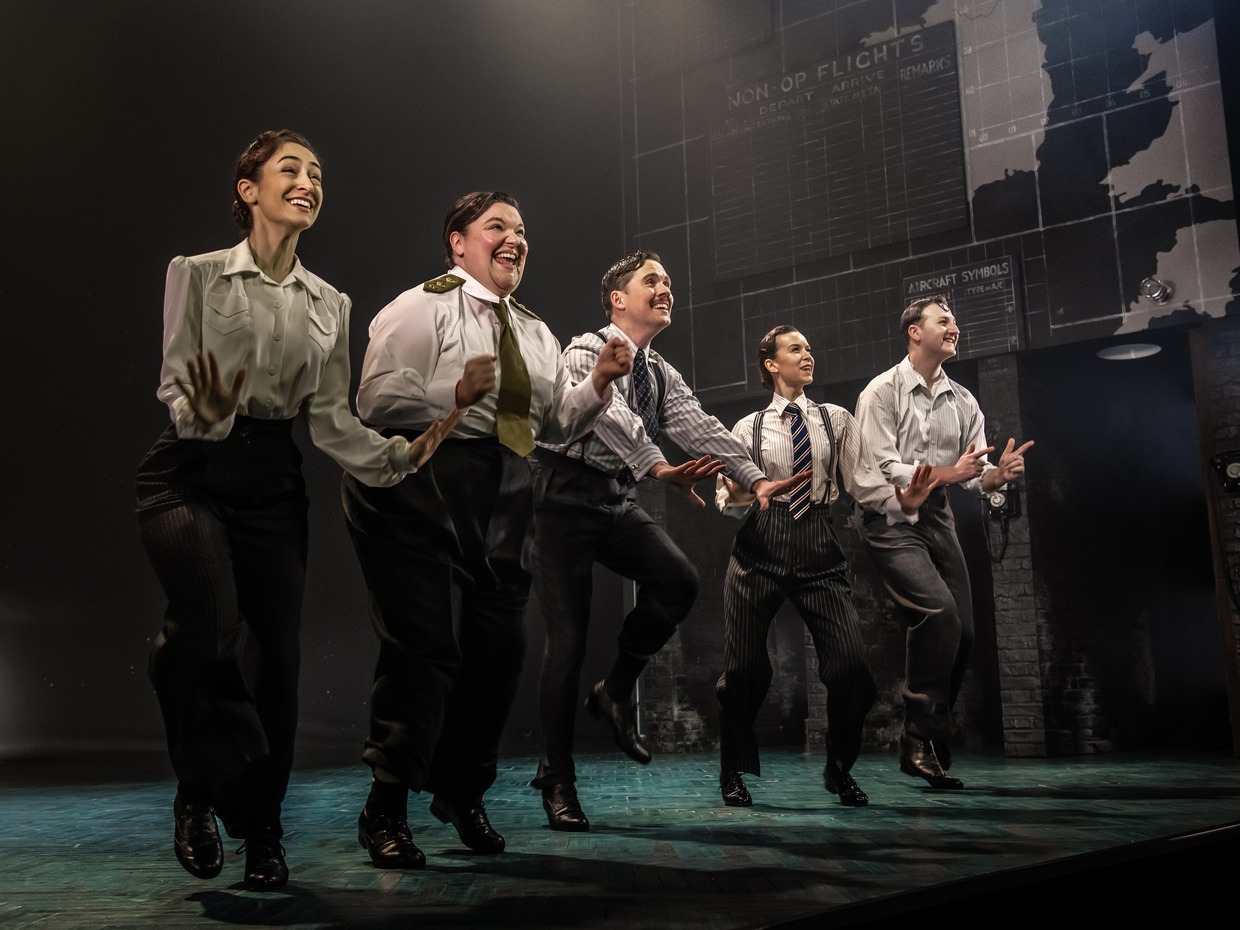 Five performers in 1940s shirts, ties and braces dancing in a line on stage in front of a wartime operations board and map
