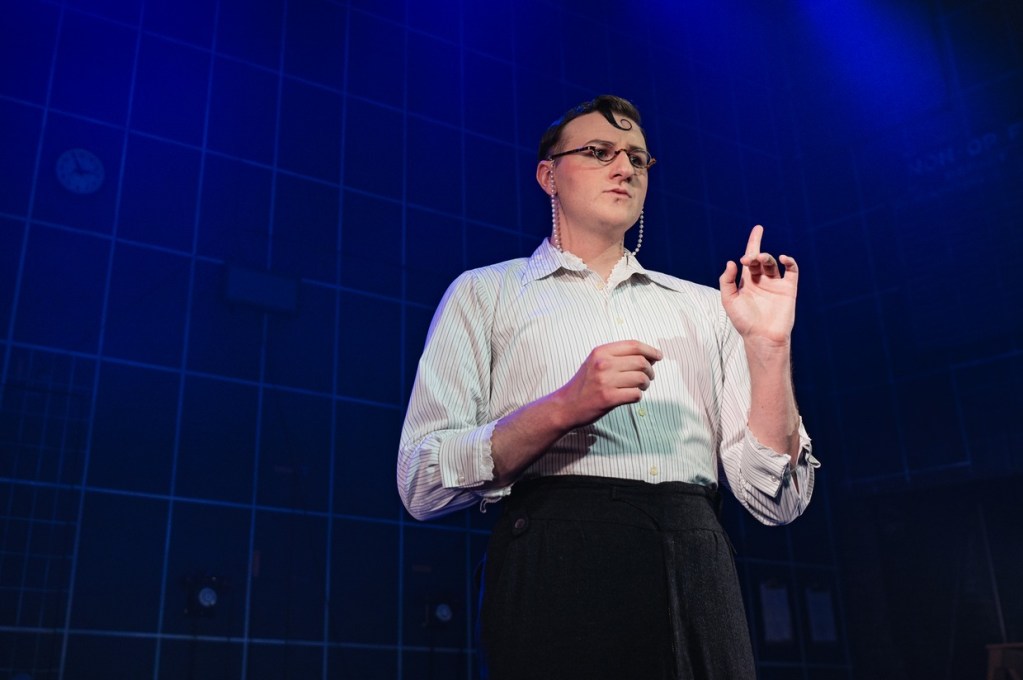 A performer in half-moon glasses on a pearl chain, a striped shirt and dark trousers, one finger raised, against a blue-lit grid backdrop