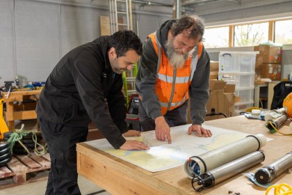 Two men studying a nautical chart of the Celtic Sea on a workbench, with cylindrical seabed recorders beside it