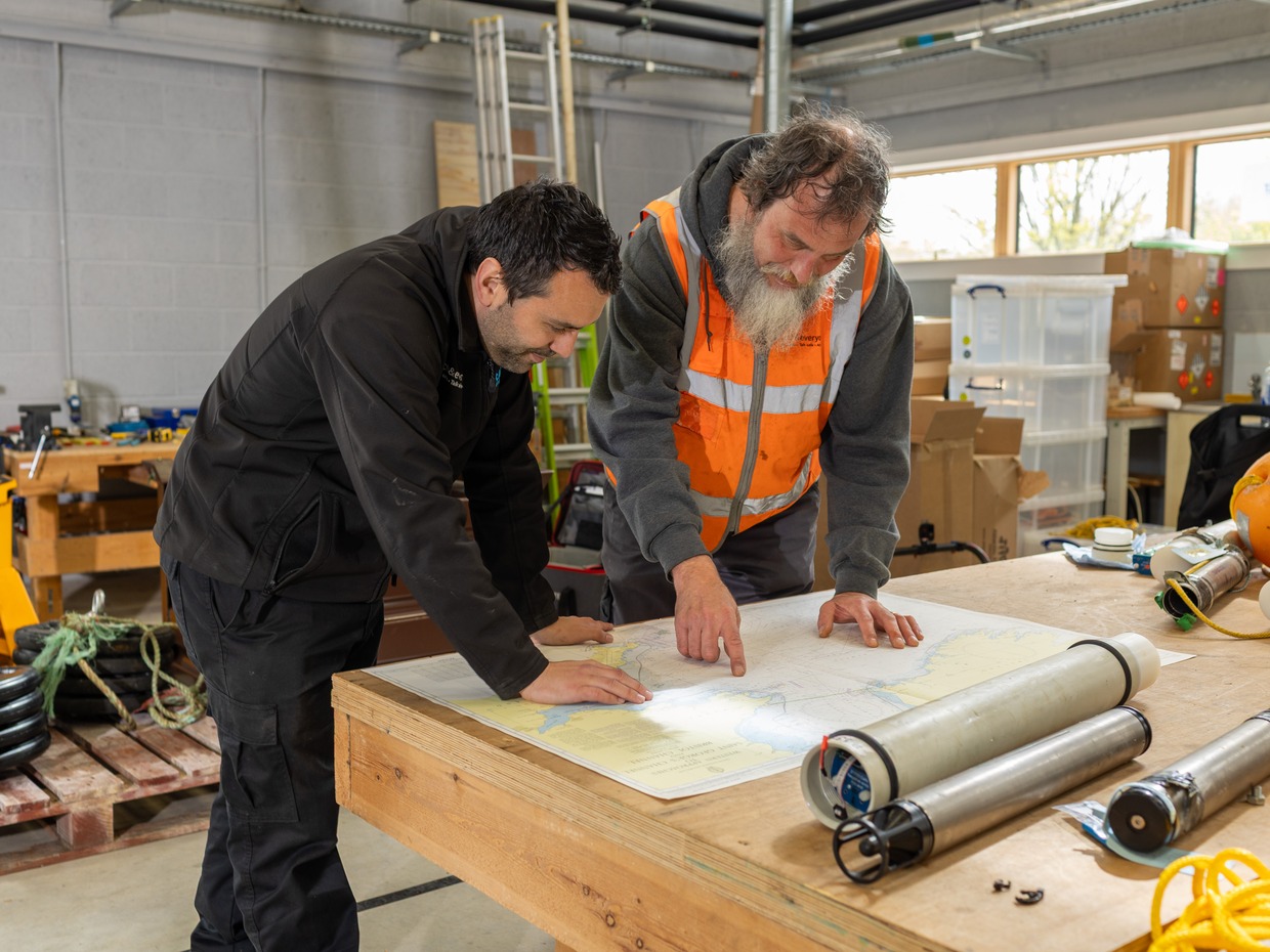 Two men studying a nautical chart of the Celtic Sea on a workbench, with cylindrical seabed recorders beside it