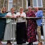 Five people leaning on a glass balcony rail outside the stone-fronted Mansion House in Swansea