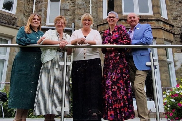 Five people leaning on a glass balcony rail outside the stone-fronted Mansion House in Swansea