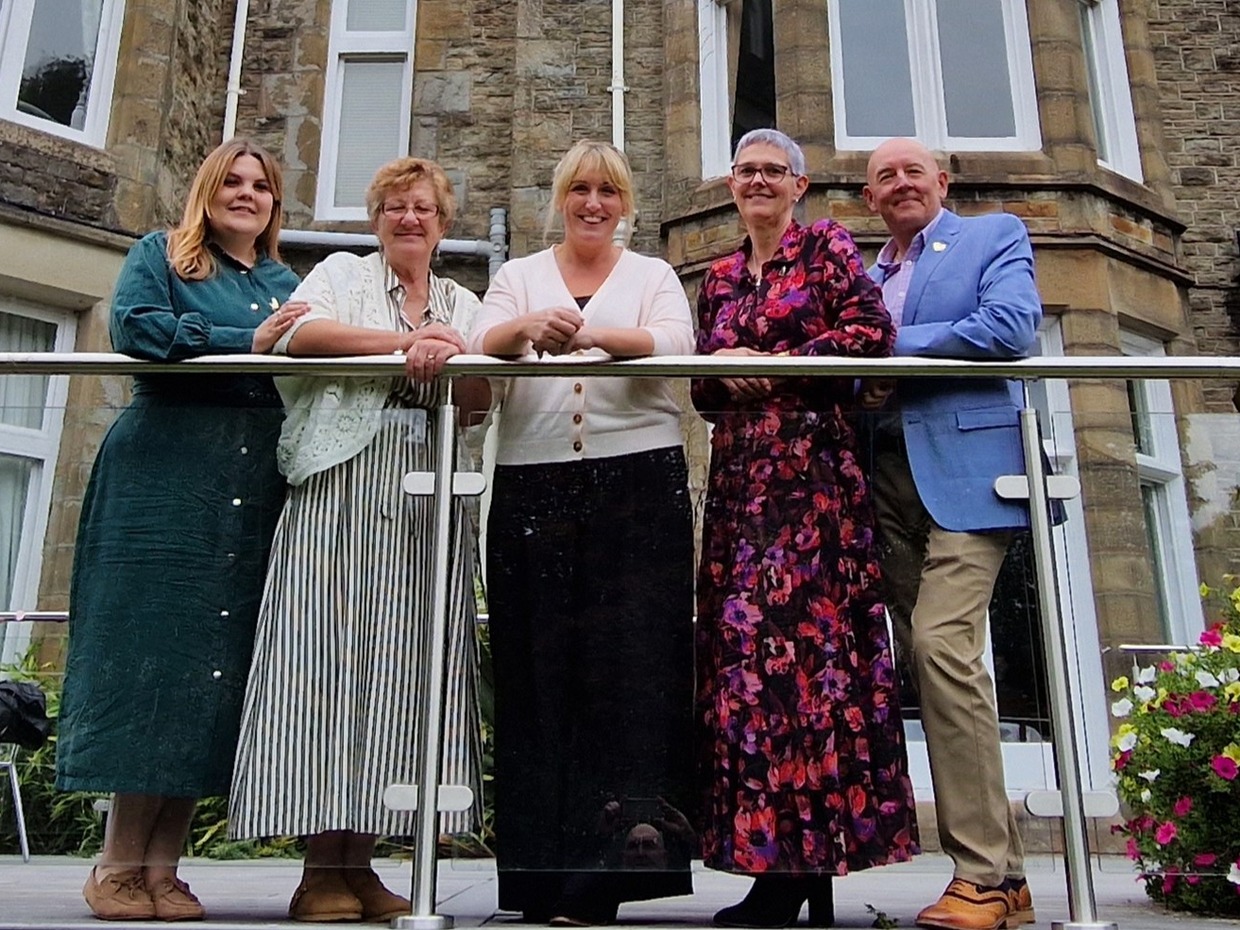 Five people leaning on a glass balcony rail outside the stone-fronted Mansion House in Swansea