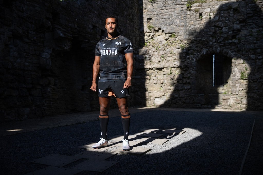 An Ospreys player in the black 2026/27 home shirt with bronze castle motifs, standing in a shaft of light inside castle ruins