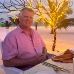 A smiling man with short grey hair in a pink short-sleeved shirt, seated at a table on a beach at dusk with fairy lights in the trees behind him