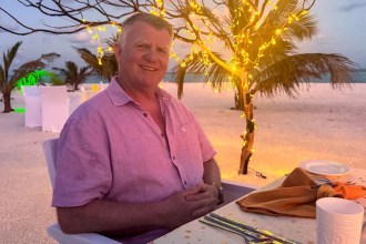 A smiling man with short grey hair in a pink short-sleeved shirt, seated at a table on a beach at dusk with fairy lights in the trees behind him