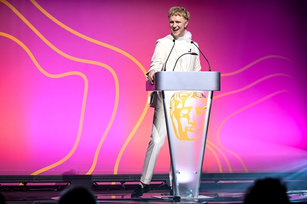 Owain Wyn Evans in a white suit smiling at a lectern bearing the gold BAFTA mask, against a pink and orange stage backdrop