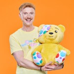 Owain Wyn Evans, smiling in a yellow Children in Need T-shirt, holding a large Pudsey Bear against an orange background