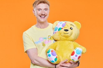 Owain Wyn Evans, smiling in a yellow Children in Need T-shirt, holding a large Pudsey Bear against an orange background