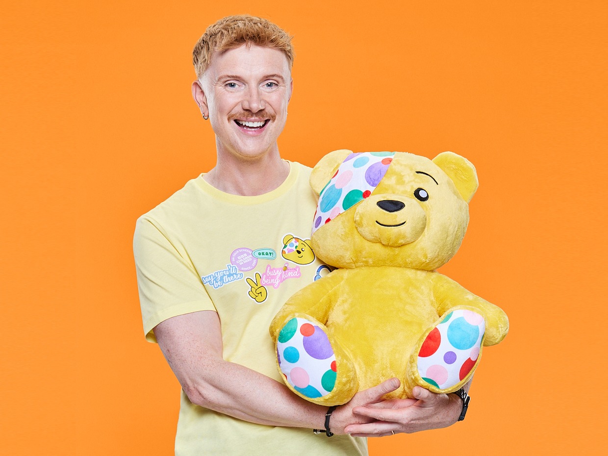 Owain Wyn Evans, smiling in a yellow Children in Need T-shirt, holding a large Pudsey Bear against an orange background