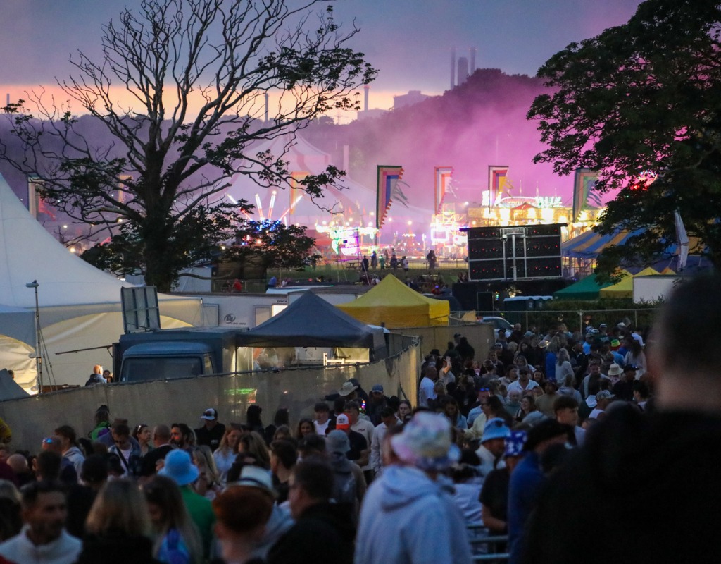 A large festival crowd walking between marquees at dusk, with colourful flags, funfair lights and pink smoke in the distance