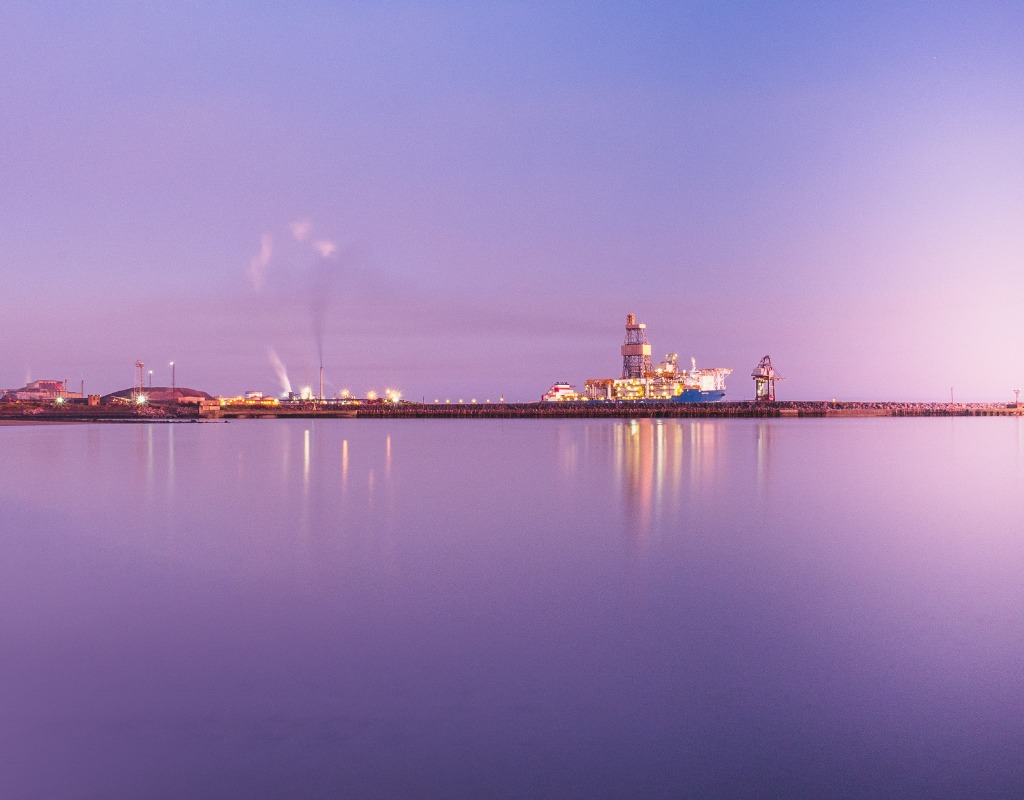 A still harbour at dusk under a purple sky, with a lit drilling rig and cranes along the breakwater reflected in the water