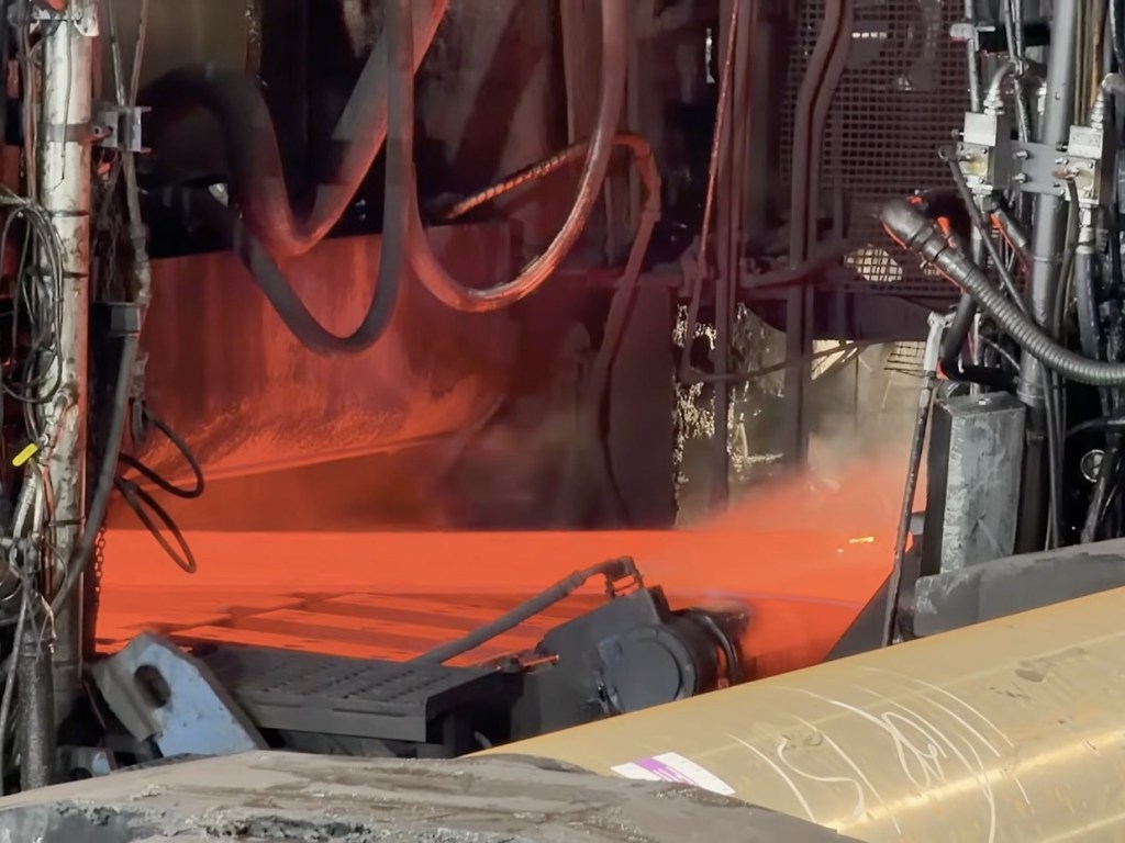 A glowing red-hot steel slab passing through the rollers of the hot strip mill, with steam and machinery around it