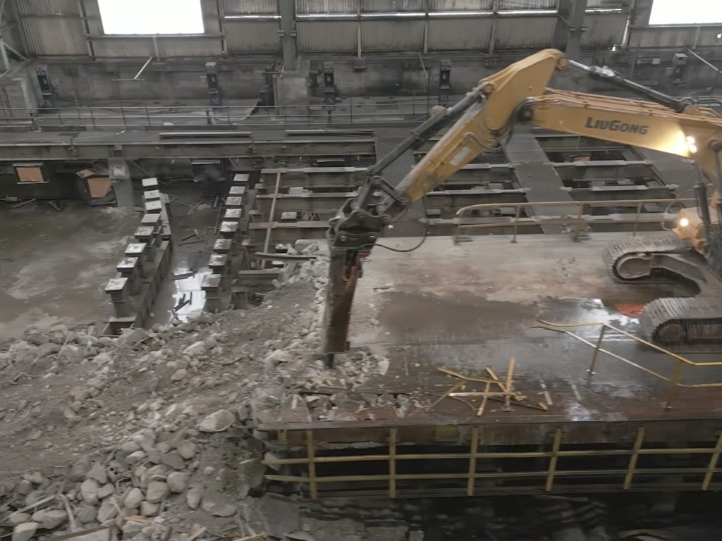 An excavator with a breaker attachment smashing concrete on the floor of the steelworks bay, with rubble piled around it