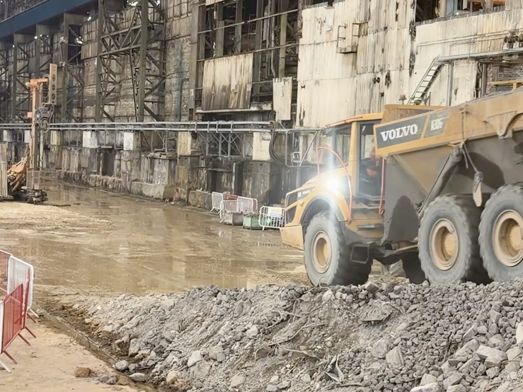 A yellow Volvo dumper truck driving across rubble inside the steelworks bay, with the old brick walls and roof structure behind