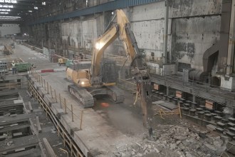 A yellow excavator working on a demolished concrete floor inside a vast steelworks bay, with rubble, exposed foundations and roof lights above
