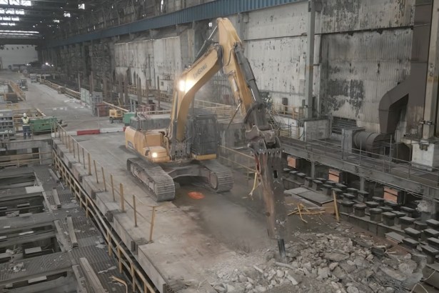 A yellow excavator working on a demolished concrete floor inside a vast steelworks bay, with rubble, exposed foundations and roof lights above