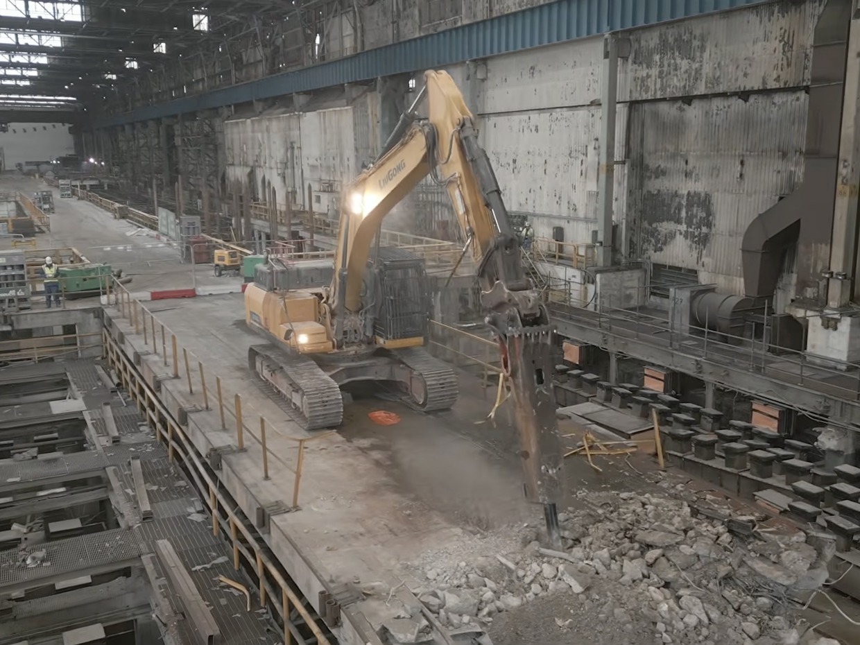 A yellow excavator working on a demolished concrete floor inside a vast steelworks bay, with rubble, exposed foundations and roof lights above