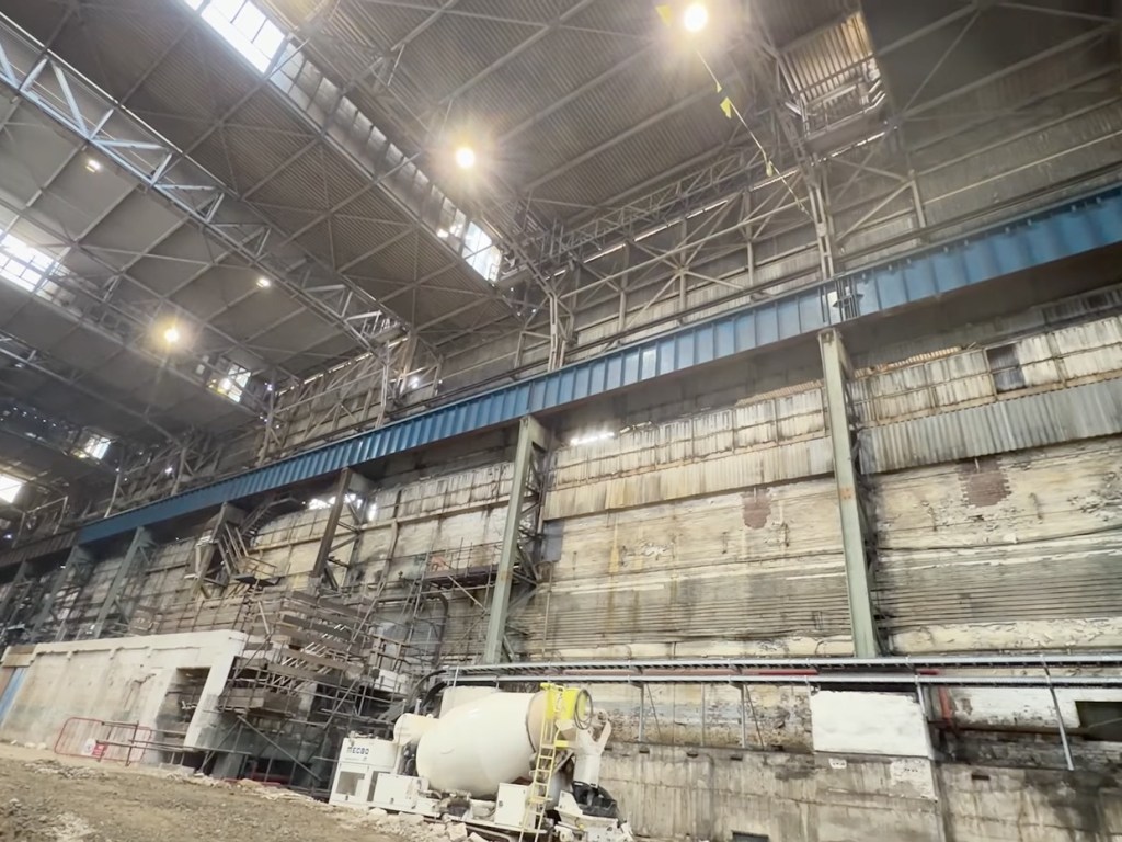 Looking up inside the cavernous steelworks bay at the roof trusses, crane gantry rails and side walls, with machinery on the floor below