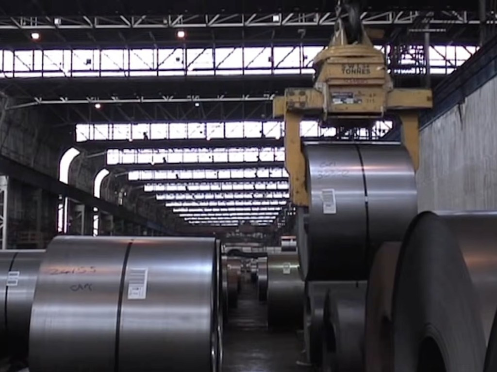 Large steel coils stacked on the floor of a steelworks bay with an overhead crane hook above them and roof lights beyond
