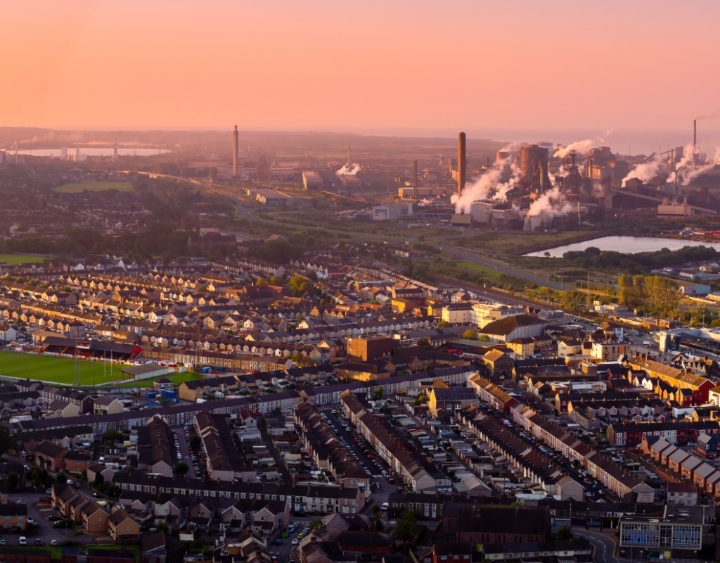 Rows of terraced houses and a rugby pitch in the foreground with the steelworks’ chimneys and steam under an orange sunrise sky
