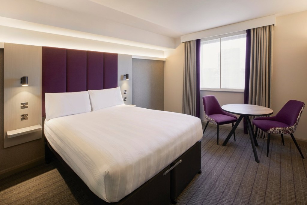 A Premier Inn double room with a purple padded headboard, white bedding, purple chairs and a small round table by the window