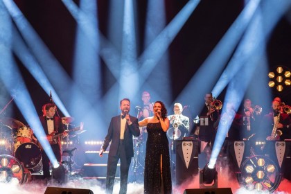 A man in a dinner jacket and a woman in a black sequinned gown sing together on a smoky stage under crossing blue spotlights, with a big band in bow ties behind them