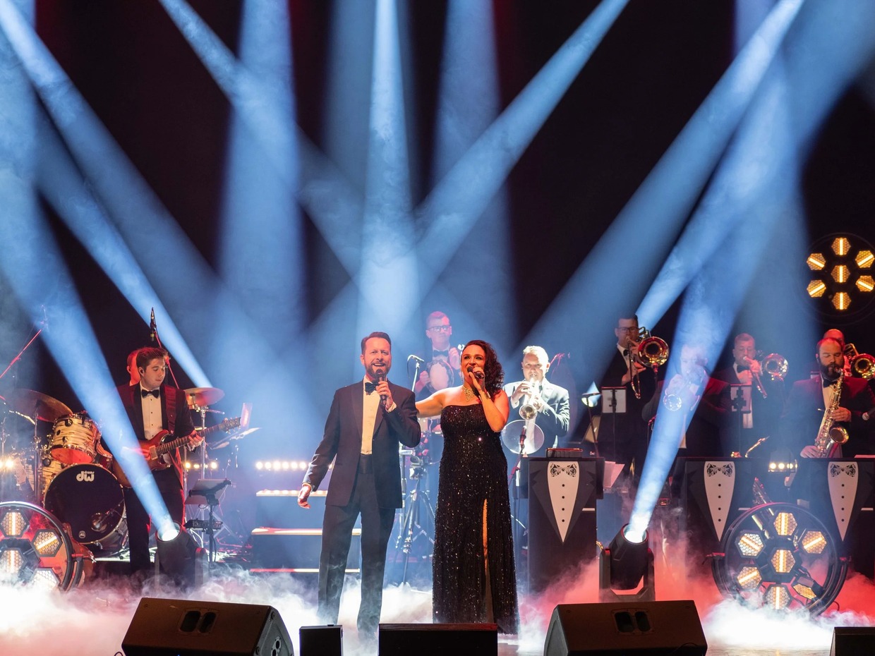 A man in a dinner jacket and a woman in a black sequinned gown sing together on a smoky stage under crossing blue spotlights, with a big band in bow ties behind them