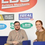 Ben and Rachel Davies sitting on blue sofas in front of a wall of LBS Group brand logos including Talbot Timber, Total Plumbing and LBS Renewables