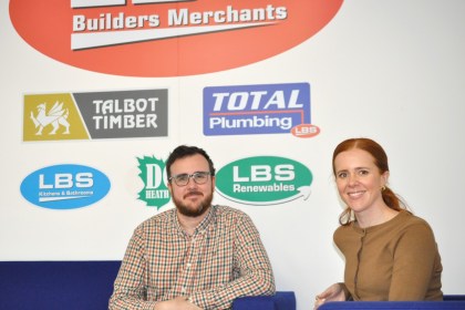 Ben and Rachel Davies sitting on blue sofas in front of a wall of LBS Group brand logos including Talbot Timber, Total Plumbing and LBS Renewables